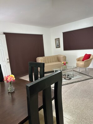 Un salon avec un canapé beige, une table basse en verre, une table à manger en bois foncé agrémentée d’un vase de fleurs et un coussin rouge sur une chaise.