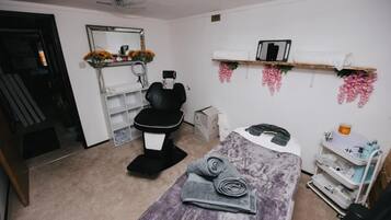 Una habitación con una camilla de masaje, una silla, un estante con toallas, un espejo y un escritorio pequeño con un teléfono y otros objetos.