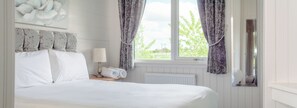 A bedroom with a bed, a nightstand, a lamp, a window with curtains, and a wall-mounted mirror.