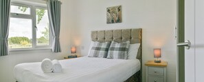 Una cama bien tendida con toallas enrolladas, un cabecero, dos mesitas de noche con lámparas y un alfeizar con cortinas.