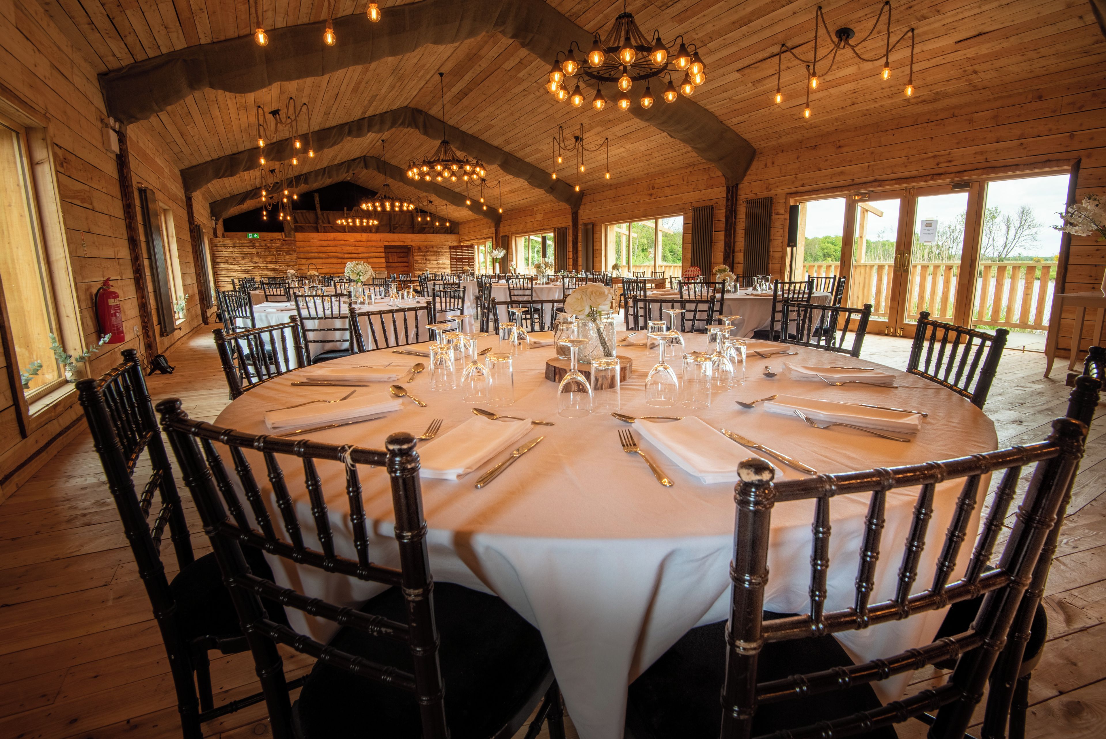 A large round table set for dining with chairs, wooden walls, and a chandelier.