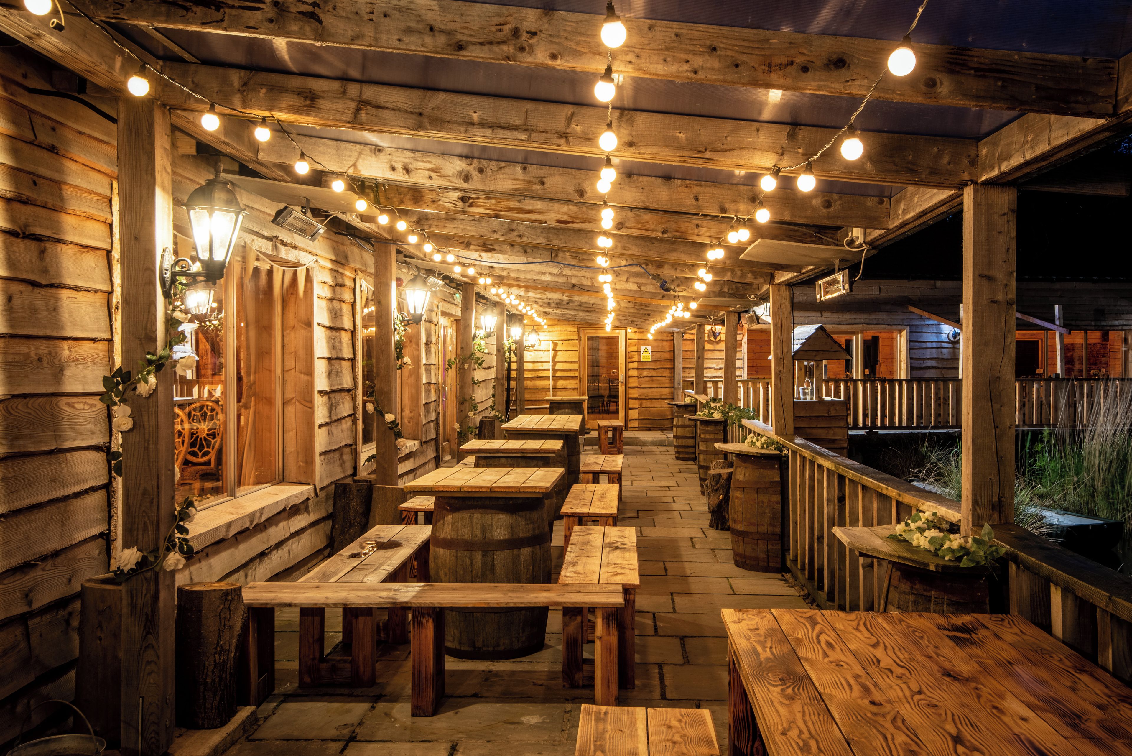 Une terrasse extérieure en bois, avec des tables et des bancs, des guirlandes lumineuses suspendues au-dessus, et un bâtiment en bois en arrière-plan.