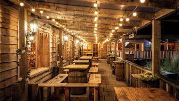 Une terrasse extérieure en bois, avec des tables et des bancs, des guirlandes lumineuses suspendues au-dessus, et un bâtiment en bois en arrière-plan.