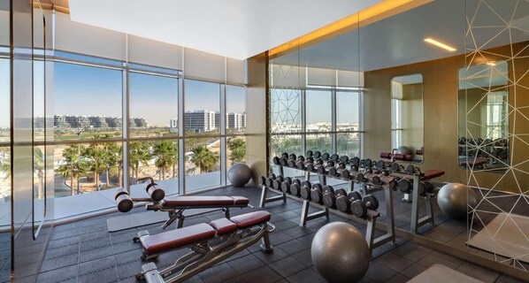 Un gimnasio con equipo de ejercicio, ventanas amplias y vistas a edificios y palmeras.
