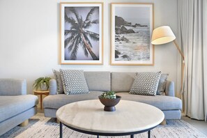 Ein Wohnzimmer mit grauem Sofa, zwei gemusterten Kissen, einem kleinen Beistelltisch mit Pflanze, einem runden Couchtisch mit Pflanze und zwei gerahmten Bildern an der Wand.