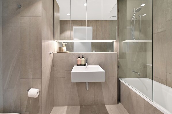 Ein modernes Badezimmer mit einer gläsernen Duschkabine, einem weißen Waschbecken mit zwei Shampooflaschen und einem Toilettenpapierhalter.