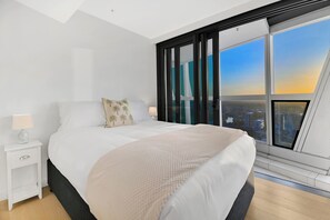A modern bedroom with a large bed, a nightstand, a lamp, and a view of a cityscape through a large window. - Jewel 27 Luxury 5 Star Beachfront King & Queen (Surfers Paradise)