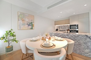 A modern dining area with a round table set for four, a kitchen with a marble countertop, and a minimalist painting on the wall. - Jewel 27 Luxury 5 Star Beachfront King & Queen (Surfers Paradise)