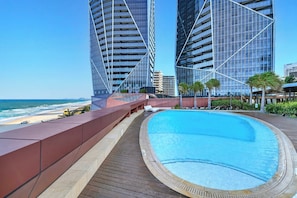 A swimming pool with a wooden deck, overlooking a beach and modern buildings. - Jewel 27 Luxury 5 Star Beachfront King & Queen (Surfers Paradise)