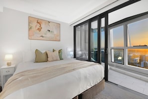 A modern bedroom with a large bed, a nightstand, a bedside lamp, a city view through a large window, and a painting on the wall. - Jewel 27 Luxury 5 Star Beachfront King & Queen (Surfers Paradise)
