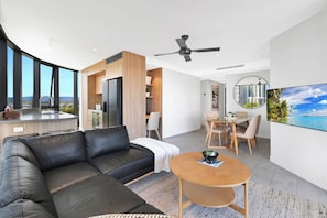 A modern living space with a black leather sofa, a round wooden coffee table, a dining area with a table and chairs, and a large flat-screen TV mounted on the wall. - Tranquillity at Encore 2BR Sea & Hinterland Views (Broadbeach)