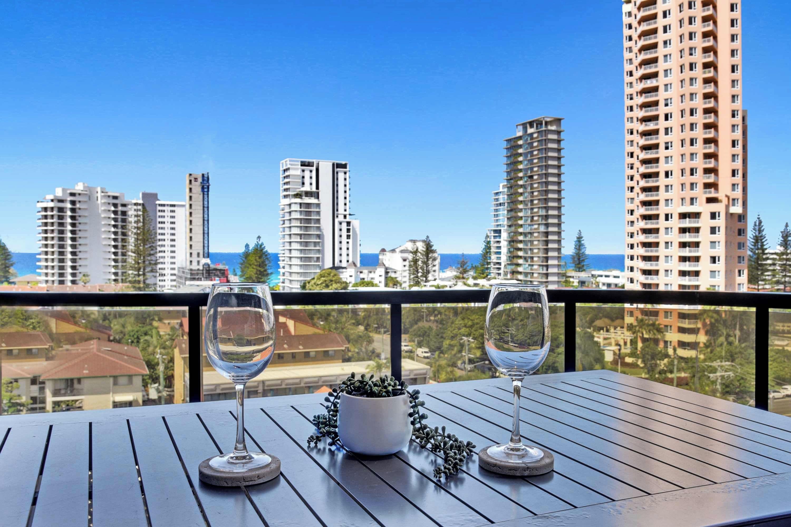A balcony with two wine glasses and a potted plant, overlooking a cityscape with tall buildings and a clear sky.