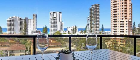 A balcony with two wine glasses and a potted plant, overlooking a cityscape with tall buildings and a clear sky.