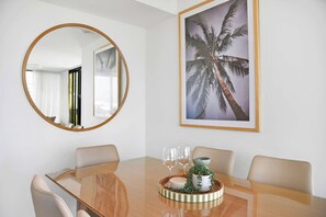 A dining area with a round table, chairs, a mirror, and a framed picture of palm trees. - Tranquillity at Encore 2BR Sea & Hinterland Views (Broadbeach)