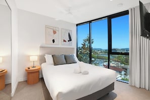 A modern hotel room with a large bed, two bedside tables, a ceiling fan, and a city view through floor-to-ceiling windows. - Tranquillity at Encore 2BR Sea & Hinterland Views (Broadbeach)