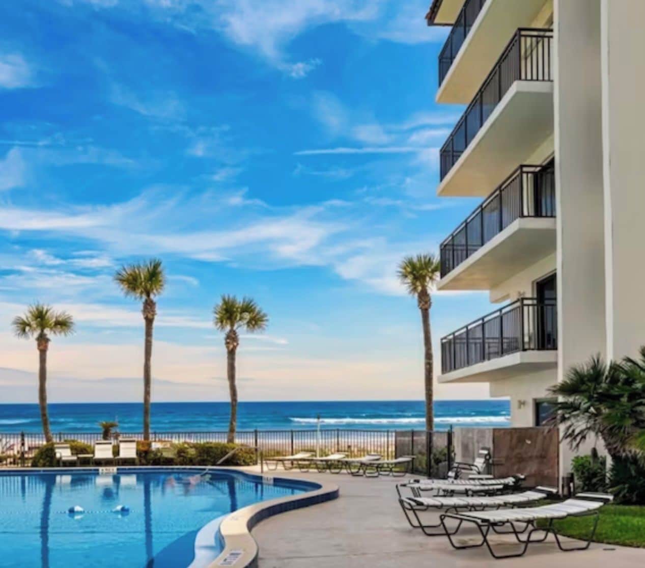 A pool area with a view of the ocean, palm trees, and a building with balconies.