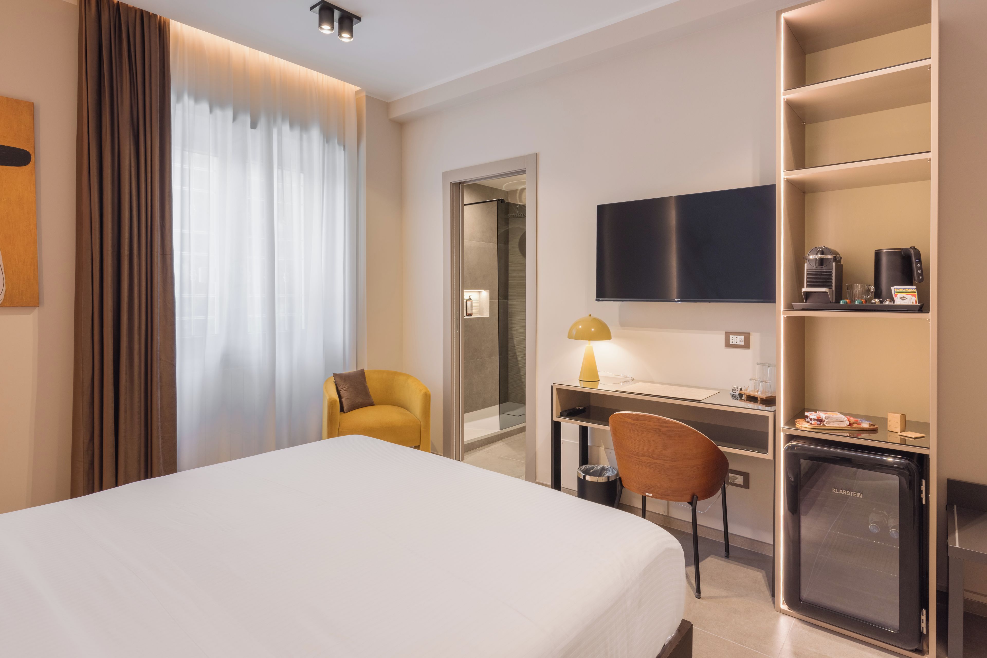 Ett hotellrum med en säng, ett skrivbord, en stol, en TV och en minibar.