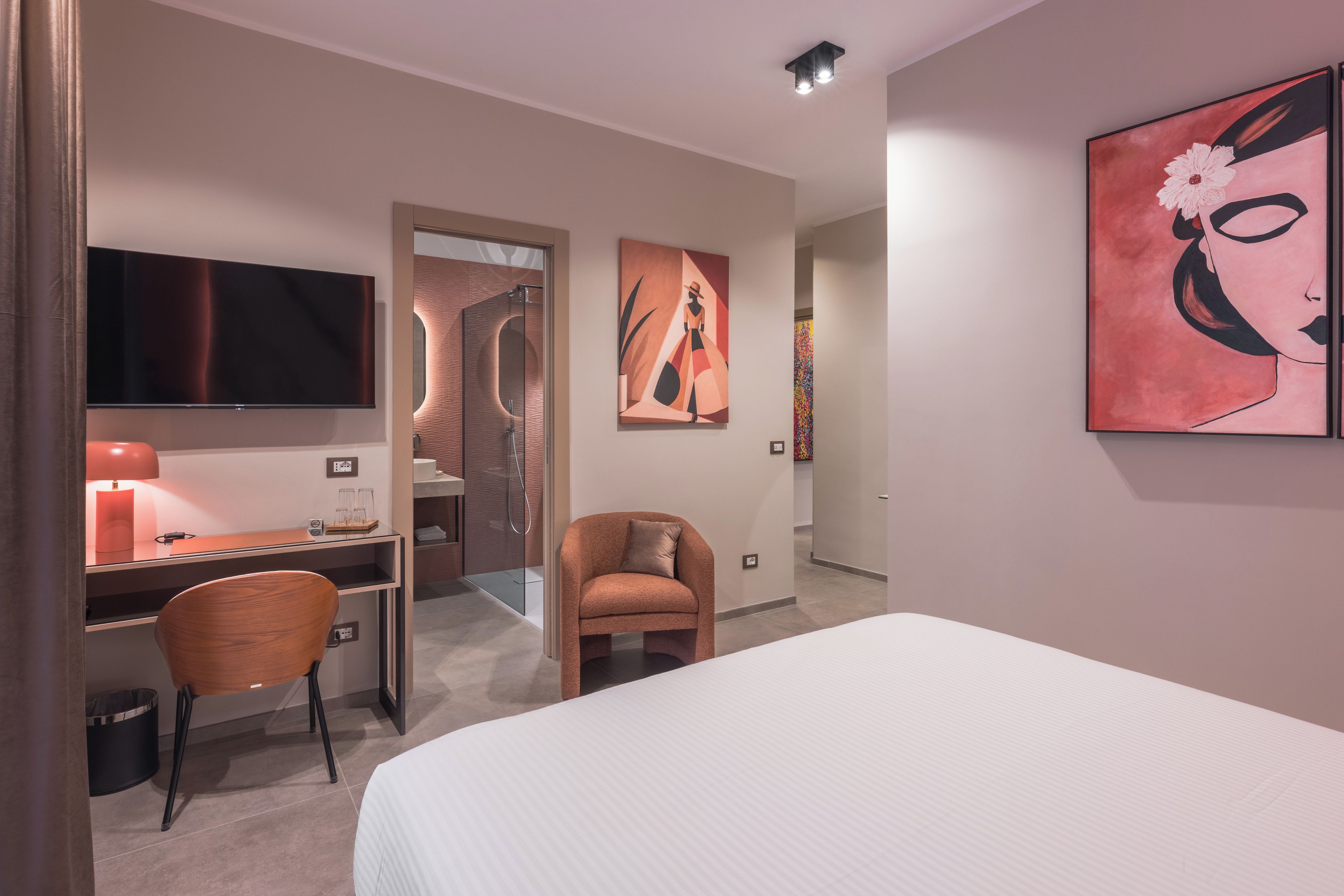 A modern hotel room with a bed, a desk with a computer, a chair, a TV, and artwork on the walls.