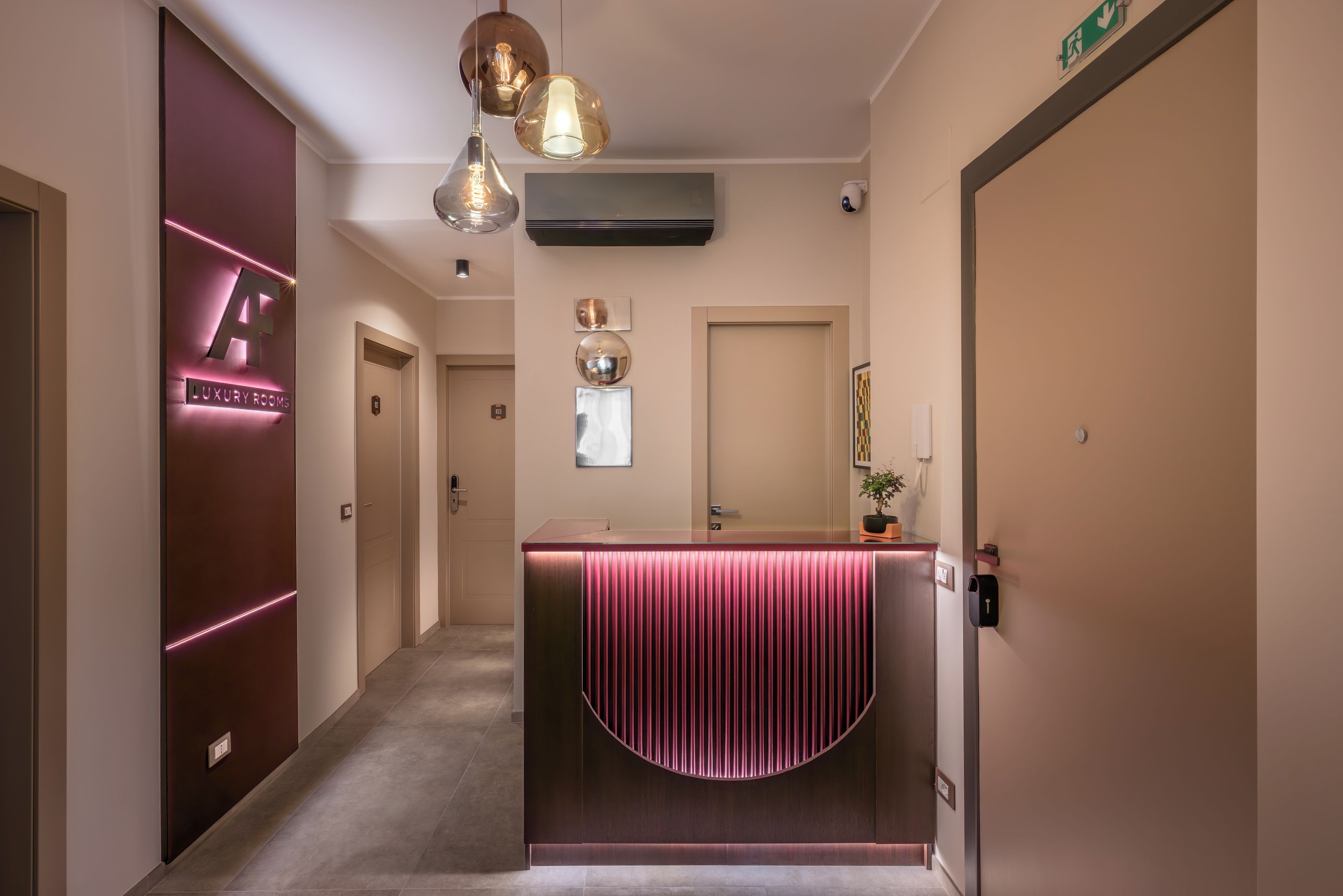 A hotel reception area with a modern design, including a reception desk with a pink accent, a digital sign indicating room types, and a security camera.