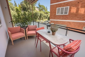 Varanda com mesa e cadeiras, um vaso de plantas e vista para estacionamento e árvores.