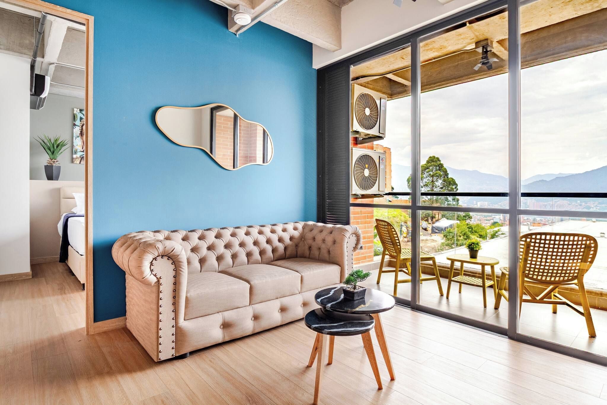 Una sala de estar moderna con un sofá color beige, una mesa redonda con una planta en maceta, y vistas de la ciudad a través de grandes ventanales.