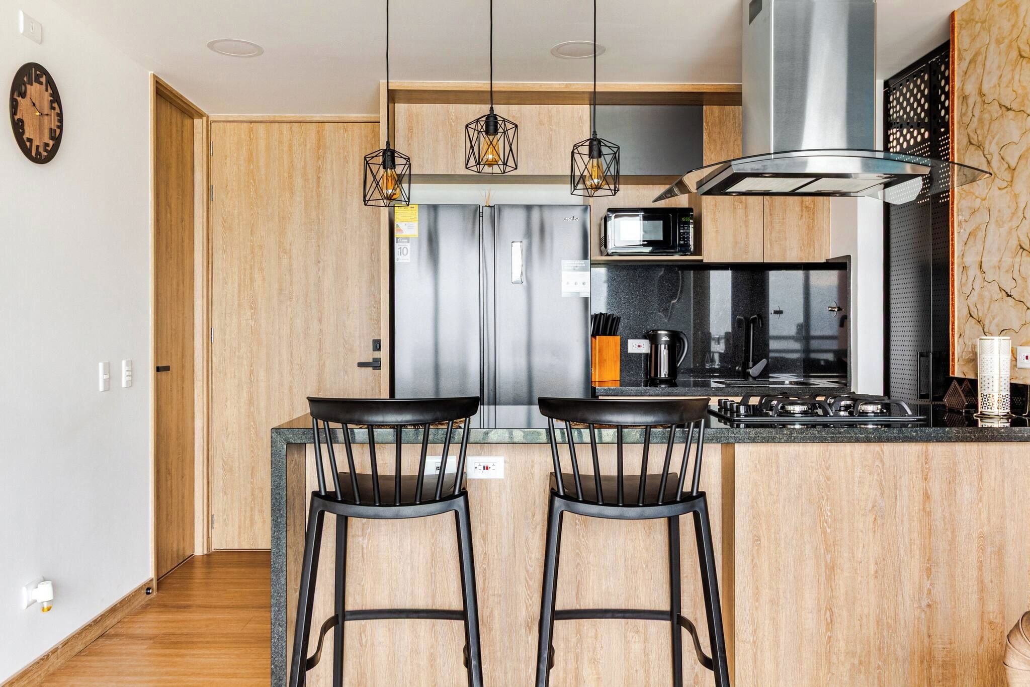 Una cocina moderna con una campana extractora de acero inoxidable, un microondas y una cafetera en la encimera. Hay dos taburetes negros frente a la encimera.