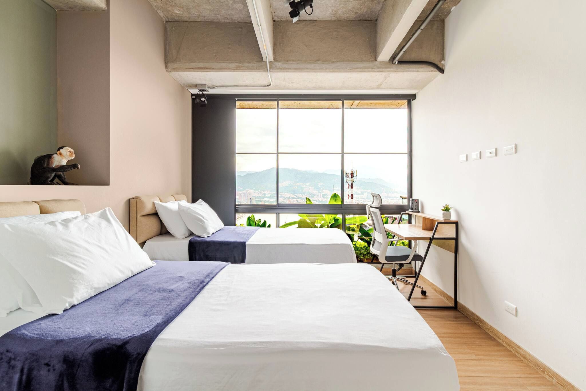 A modern bedroom with two beds, a desk, and a view of a cityscape.