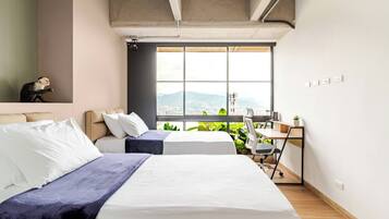 A modern bedroom with two beds, a desk, and a view of a cityscape.