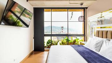 A bedroom with a bed, a TV mounted on the wall, and a large window with a city view.