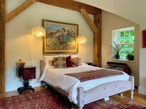 Une chambre avec un grand lit, un tableau au-dessus, des poutres en bois, une fenêtre avec des plantes et un tapis rouge.