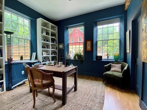 Ein Zimmer mit Schreibtisch aus Holz, einem Stuhl, einem Bücherregal, einem Fenster mit Blick auf ein rotes Gebäude und einer Zimmerpflanze.