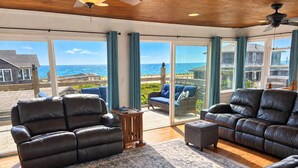 A spacious living area with leather sofas, a patterned rug, and large windows offering a view of the ocean and neighboring buildings. - Gatsby’s OBP, 3rd House to the Beach, Heated Pool + 2 Hot Tubs, Sleeps 24 (Ocean Bay Park)
