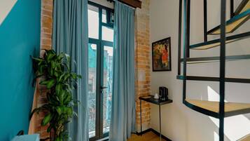 Una habitación con pared de ladrillo, ventana con cortinas, planta en maceta, cafetera y escalera de metal negro.