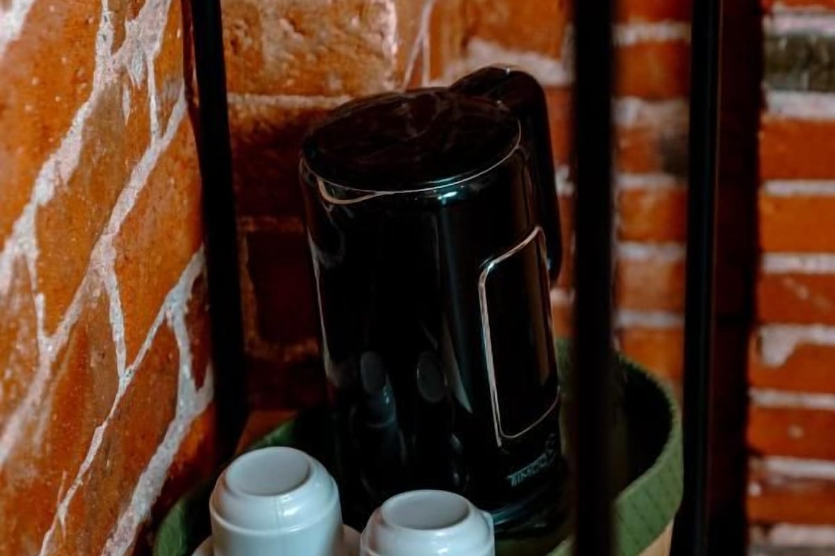 Eine Kaffeemaschine, zwei weiße Tassen und eine Zeitschrift auf einem Holztisch vor einer Ziegelwand.