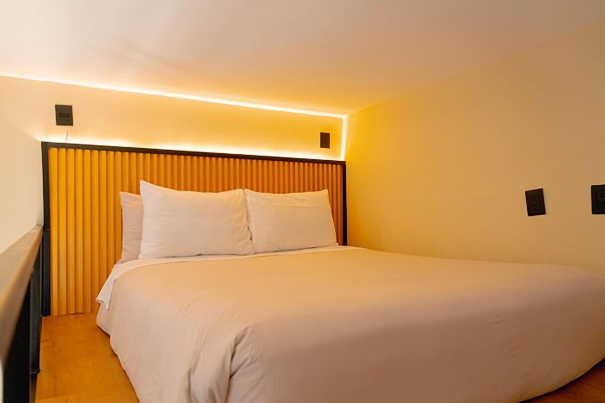 Quarto de hotel com uma cama grande, cabeceira com ripas verticais e luminária fixa na parede.