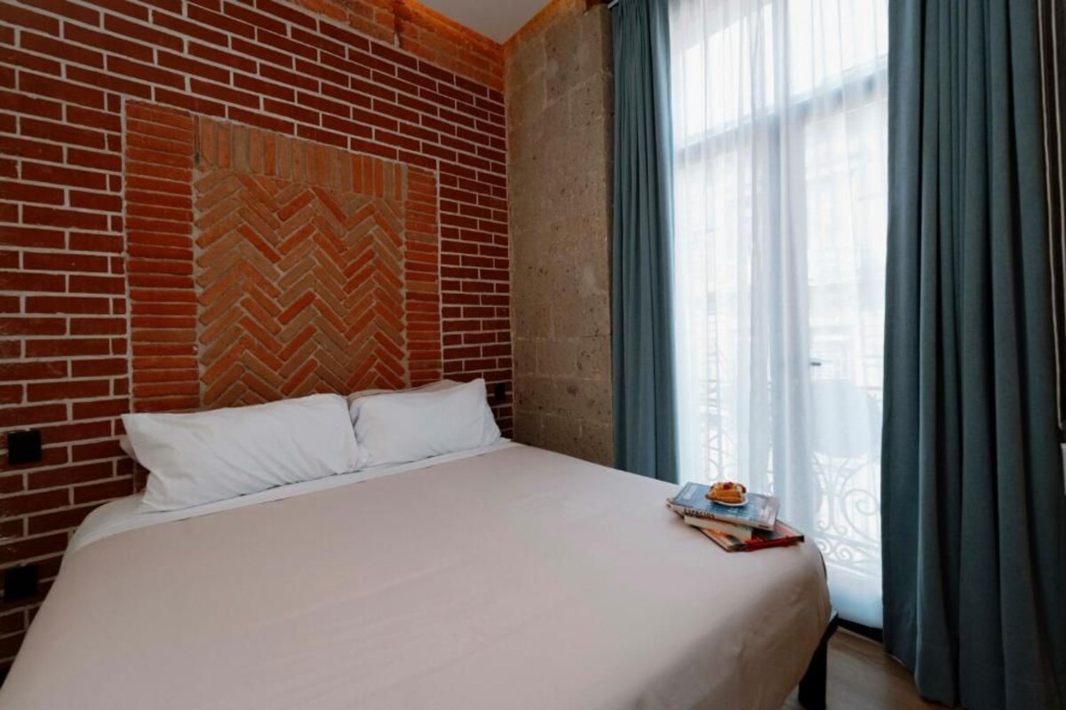 A hotel room with a brick wall, a bed with white bedding, and a window with curtains.
