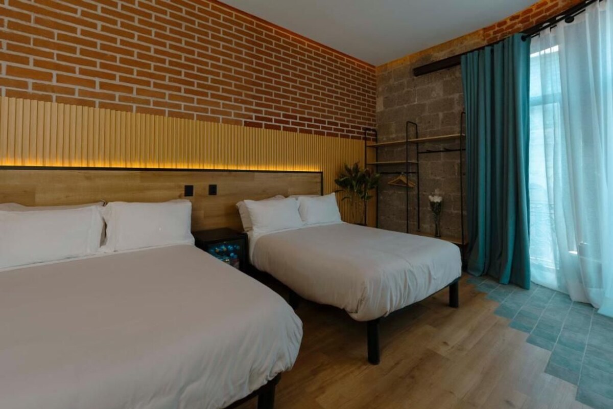 A hotel room with two beds, a brick wall, a wooden headboard, a window with curtains, and a plant.