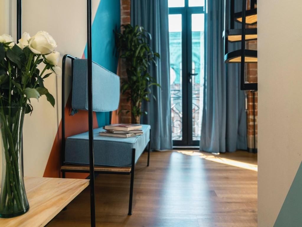 A modern room with a wooden floor, a blue sofa, a small table with a vase of flowers, and a large window with curtains.