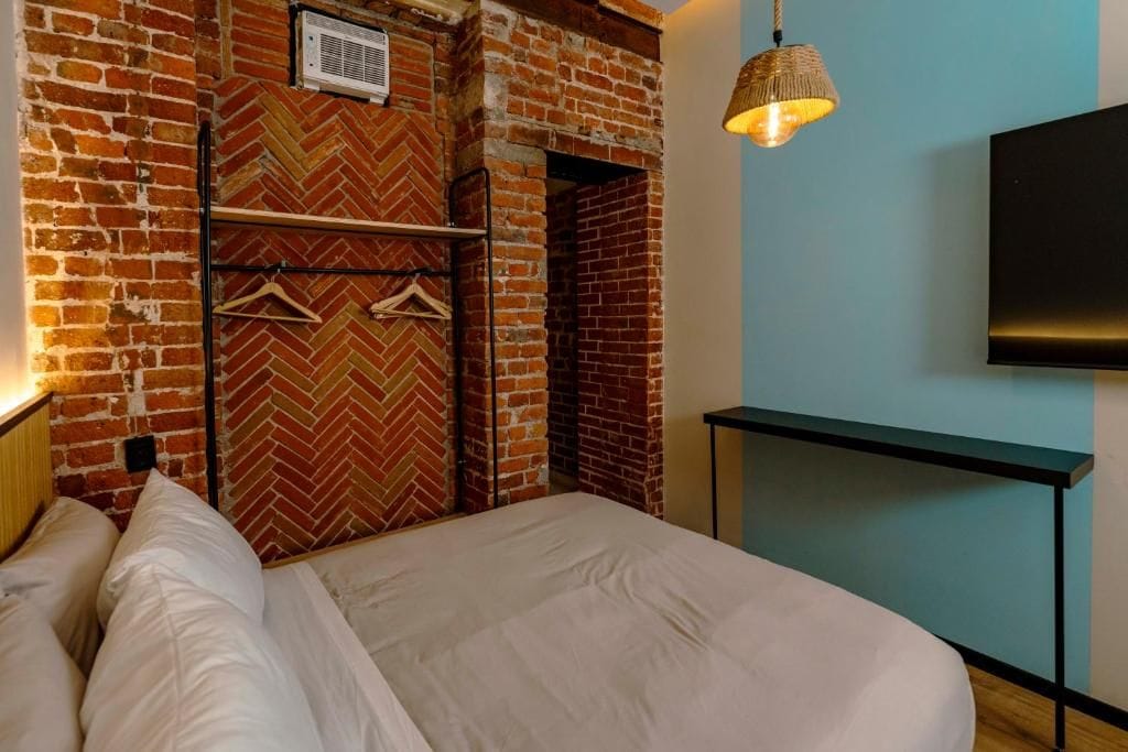 Una camera d'albergo con una parete in mattoni, un letto con biancheria bianca, un lampadario e un televisore a schermo piatto fissato al muro.
