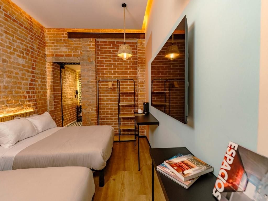 Habitación de hotel con dos camas, una pared de ladrillo, suelo de madera, un escritorio con una silla, una lámpara colgante y una revista sobre el escritorio.