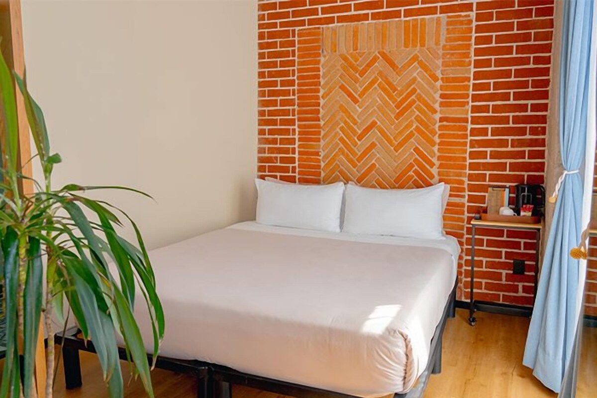 Quarto de hotel com parede de tijolos, cama com roupa de cama branca, planta e mesinha de cabeceira com abajur.