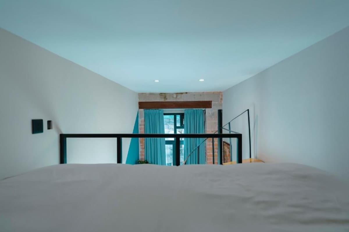 Un dormitorio con cama, ventana con cortinas, pared de ladrillo visible e iluminación empotrada en el techo.