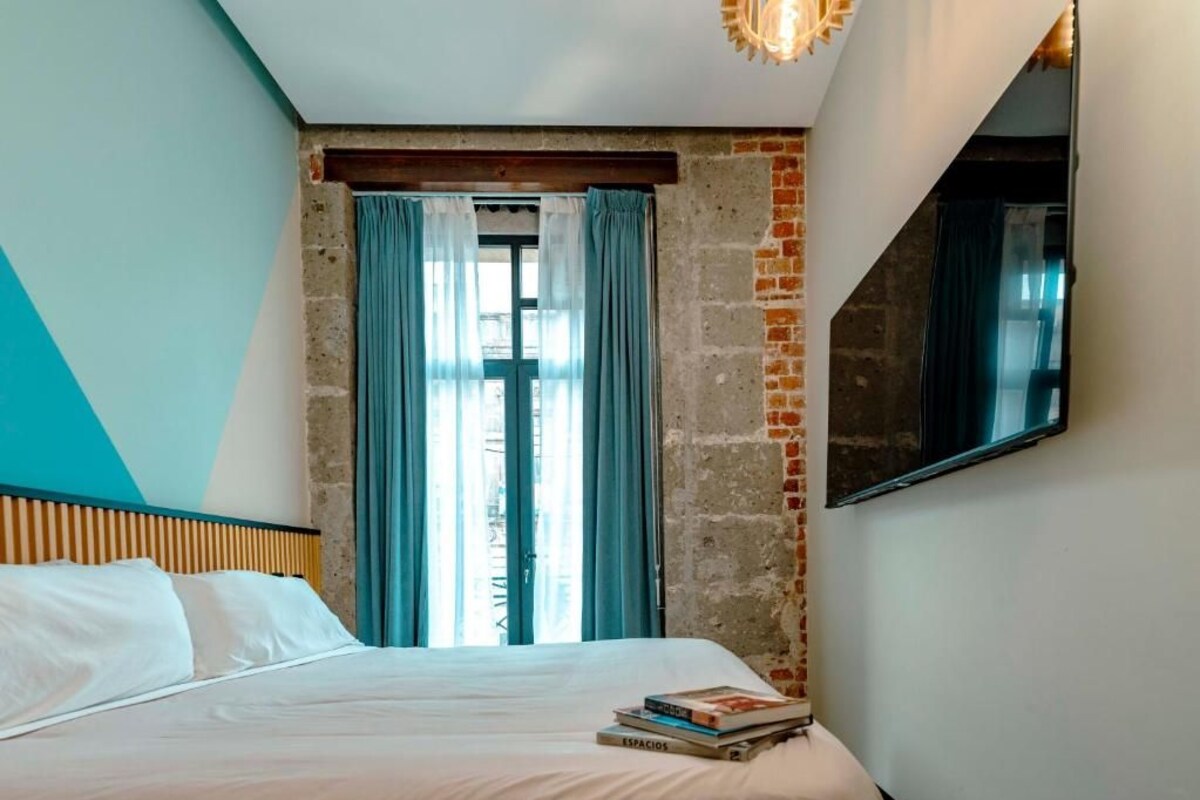 A bedroom with a bed, a window with curtains, a brick wall, and a mounted TV.