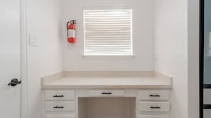 A white desk with drawers, a window with blinds, and a fire extinguisher on the wall.
