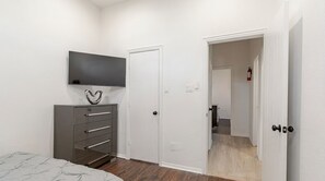 A bedroom with a bed, a dresser, a TV, and a door leading to another room.