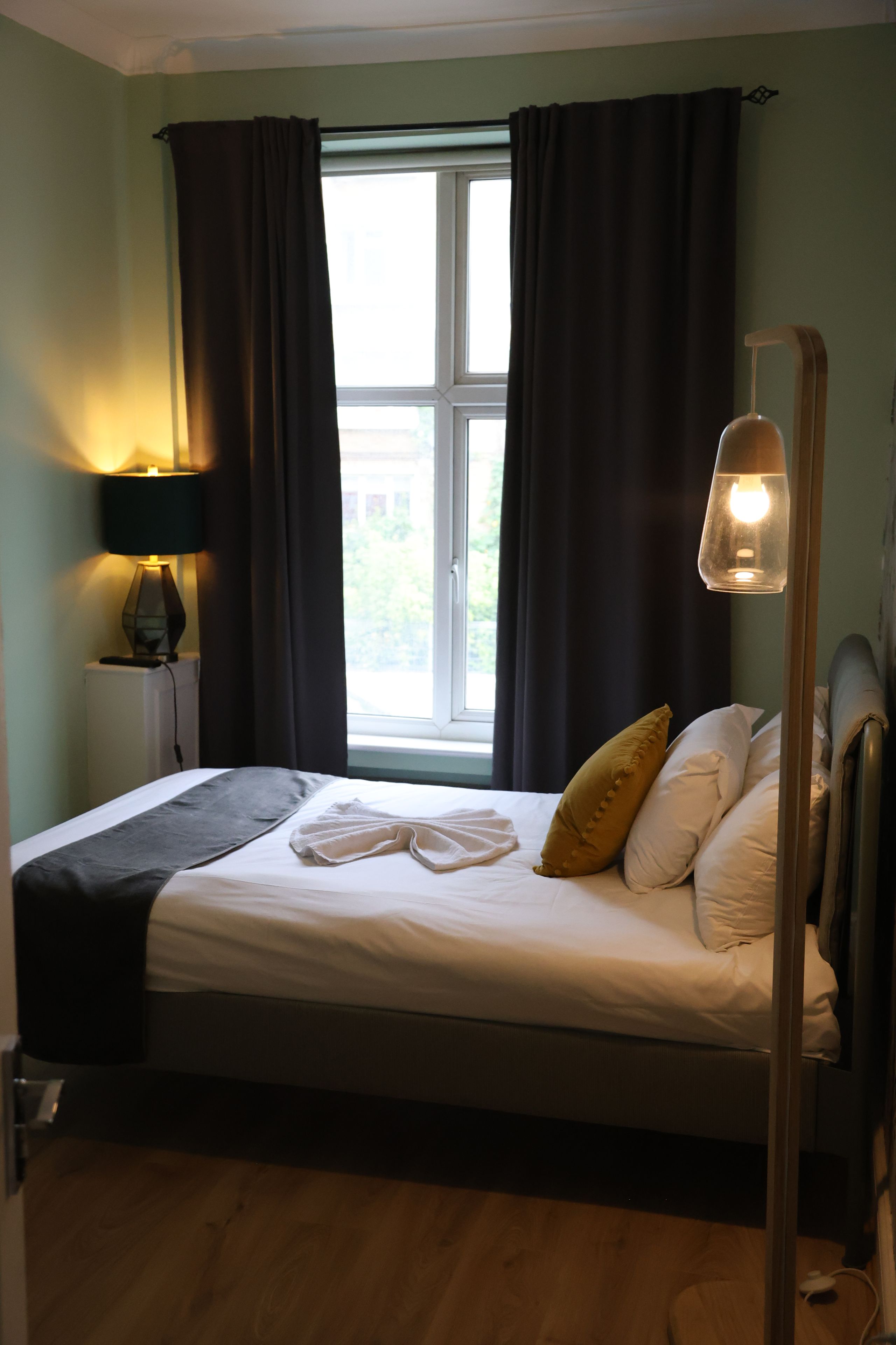 Cama bem arrumada com lençóis brancos e um único travesseiro amarelo, luminária de cabeceira e janela com cortinas.