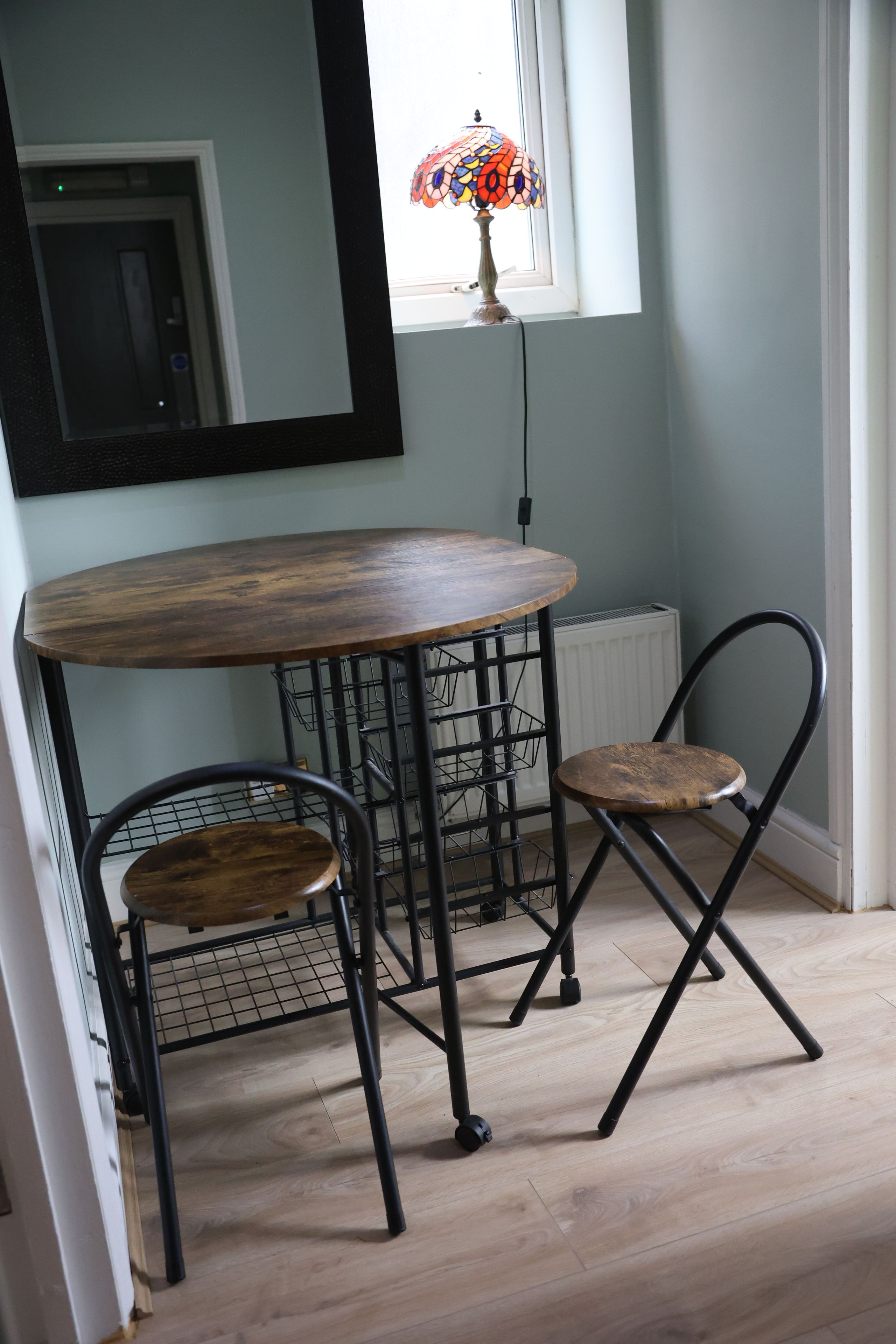 Et hjørneværelse med et rundt træbord, to metalstole, en radiator og et vindue med en farverig lampe.