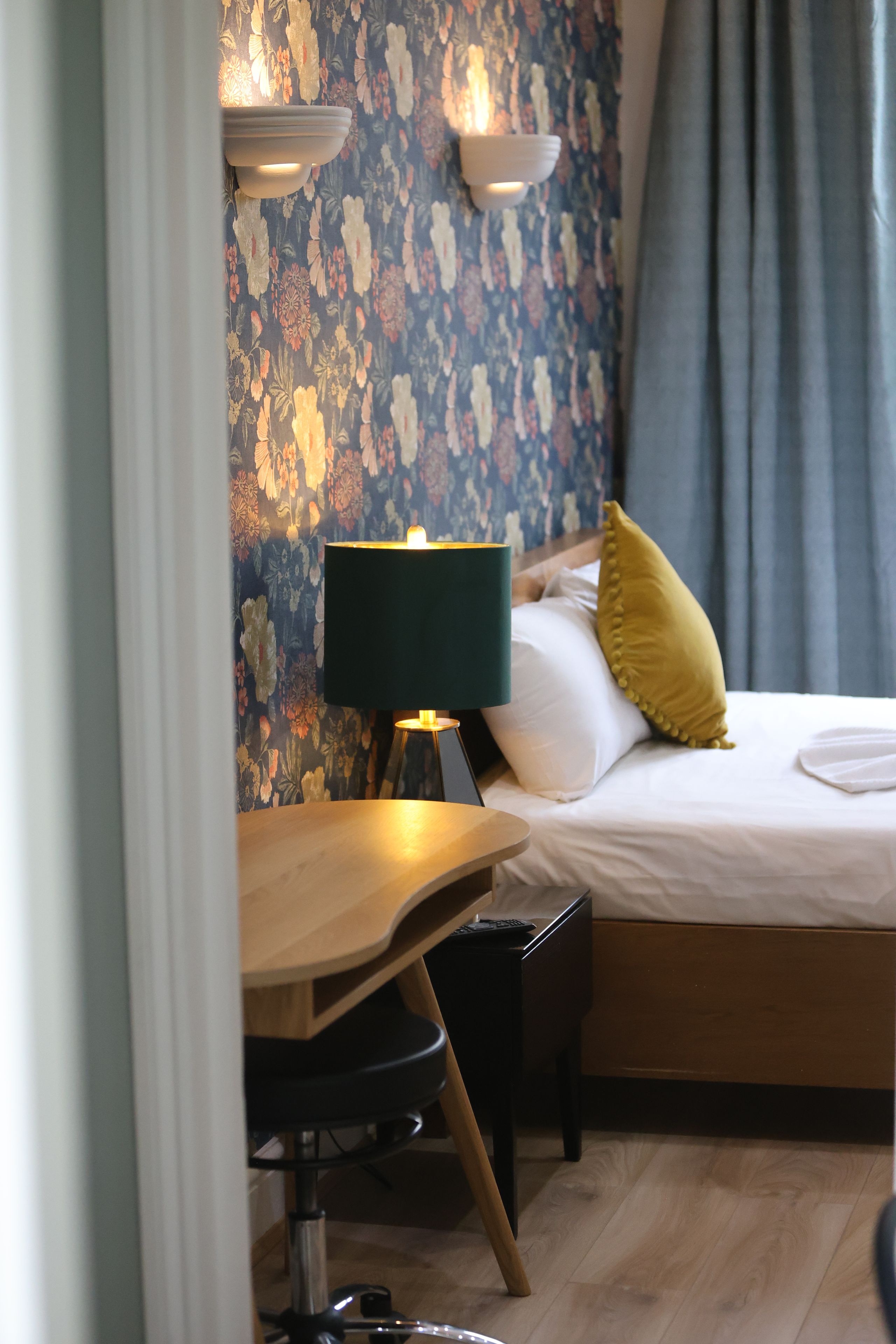 A hotel room with a bed, a desk, a lamp, and a floral wallpaper.