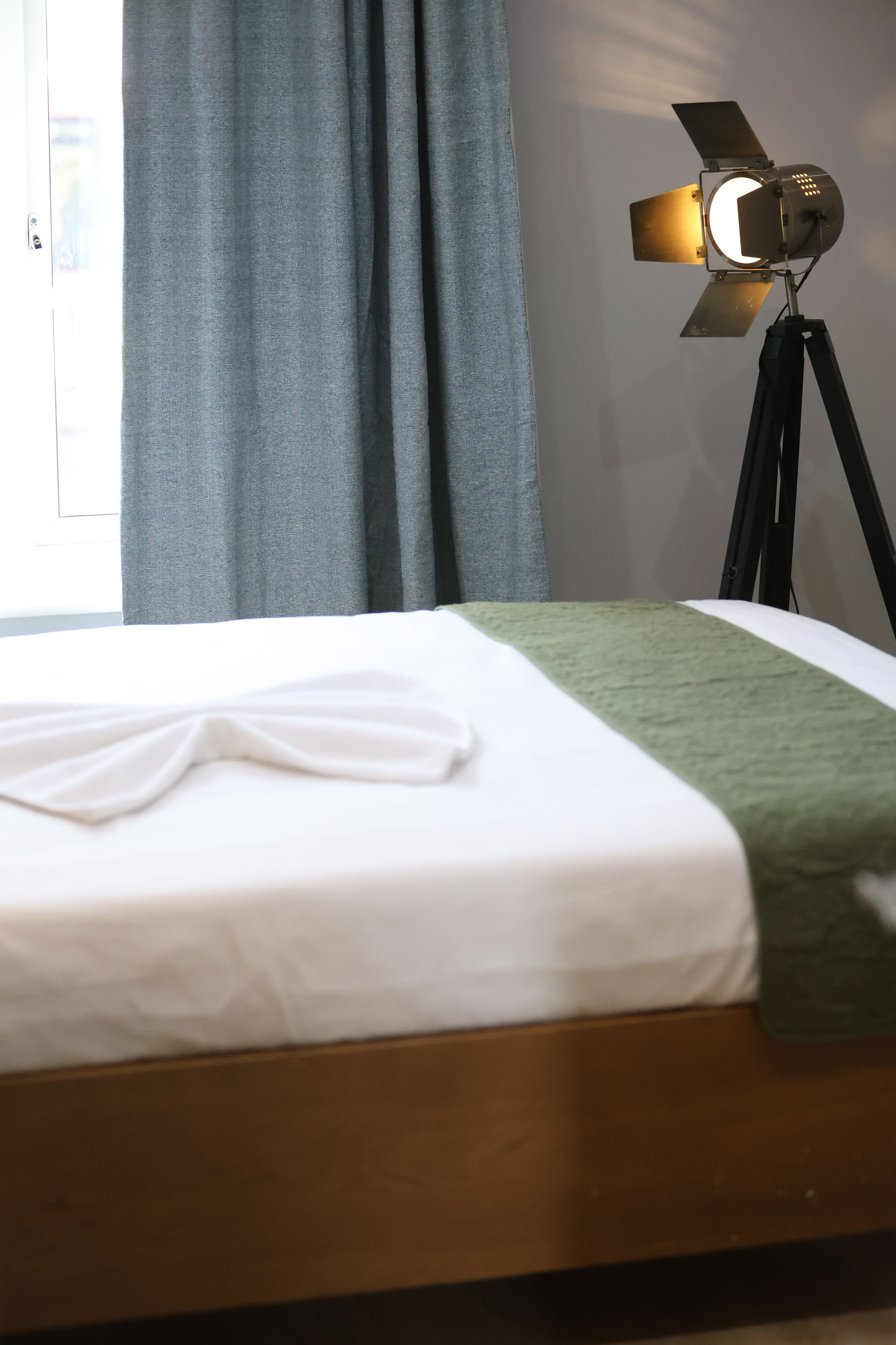 Una cama bien hecha con sábana blanca y manta verde, una lámpara sobre un trípode, y una ventana con cortinas.