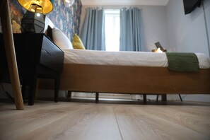 A bedroom with a bed, a nightstand, a lamp, and a window with curtains. - The Good Vibe (London)